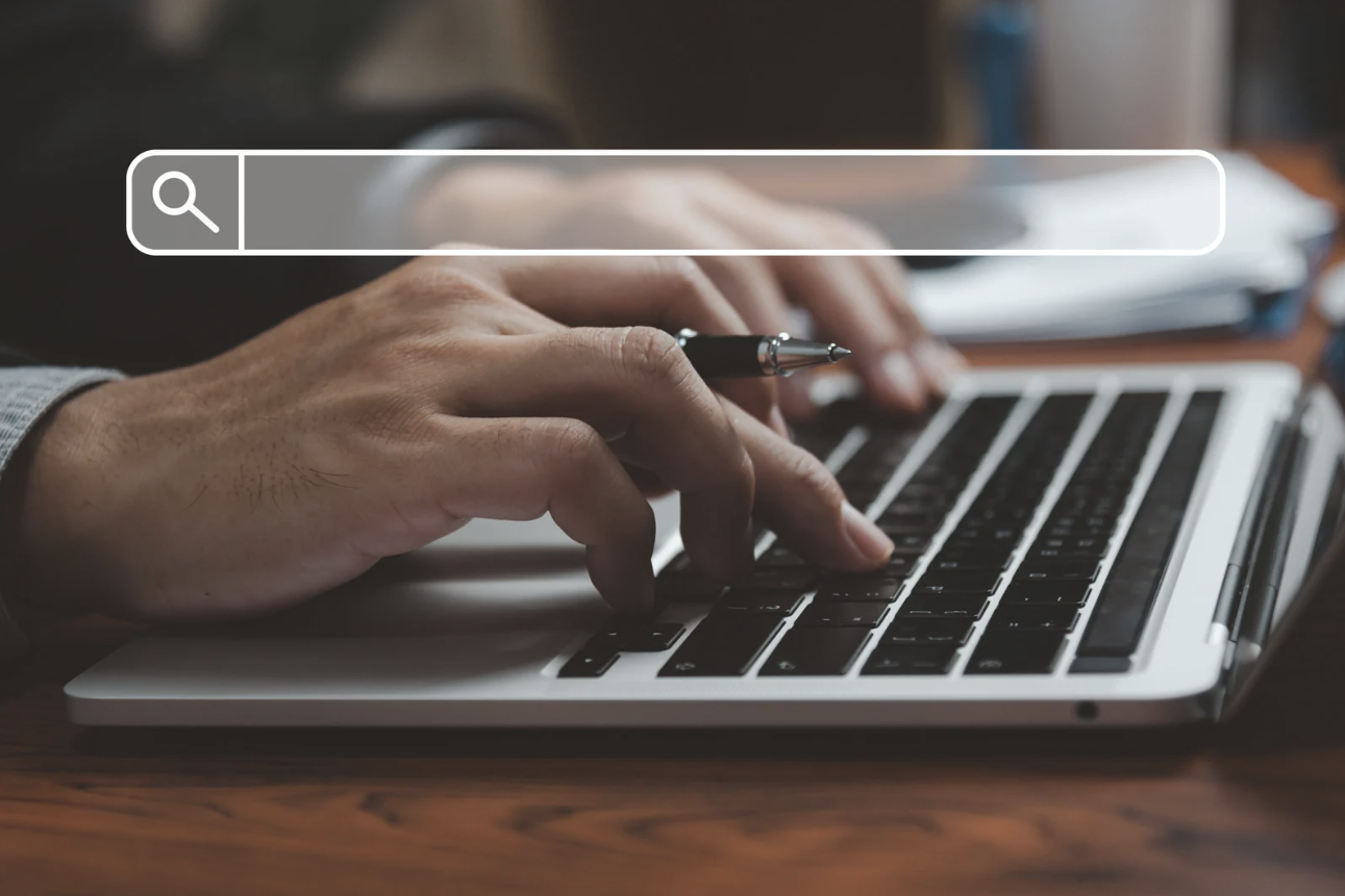 Person typing on computer while holding a pen between two fingers. A transparent search bar appears over the image.